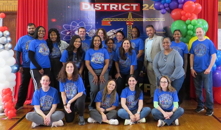 The District 24 team in blue STEM Expo shirts, gathered in a school gym under a District 24 banner and balloons